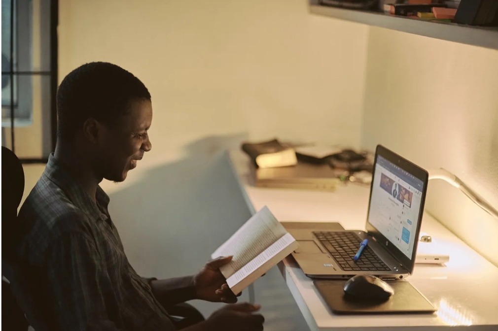 A man seated at his desk with a book and laptop